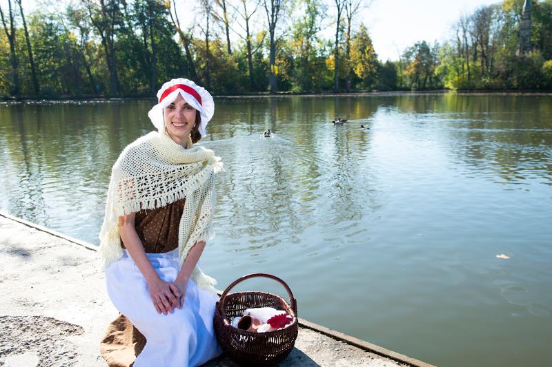 Eine Frau sitzt an einem See, sie trägt einen weißen Hut, einen weißen Schal um die Schultern und einen weißen Rock. Neben ihr steht ein geflochtener Korb.