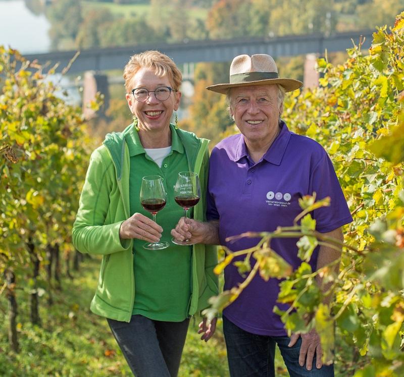 Ein Mann und eine Frau stehen zwischen Weinreben und halten jeder ein Glas Rotwein in der Hand. Sie lachen und stoßen mit den Gläsern an.