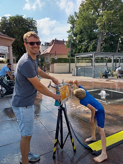 Mann füllt Flasche, Kind trinkt am Wasserspender