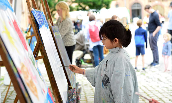 Mädchen malt an einer Staffelei. Im Hintergrund mehrere Erwachsene und Kinder.