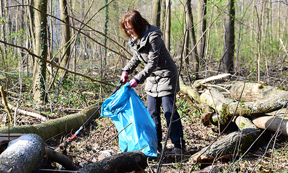 Eine Frau sammelt mit einer Zange Müll in einem Waldstück auf.