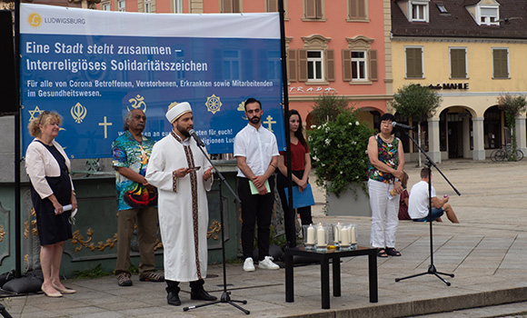 Vertreter verschiedener Religionsgemeinschaften stehen auf dem Marktplatz. Ein Mann spricht ins Mikrofon. Auf einem Banner im Hintergrund steht: Eine Stadt steht zusammen - Interreligiöses Solidaritätszeichen.