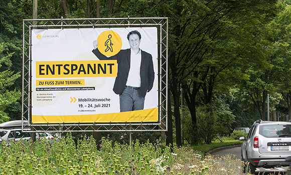 Auf der Mittelinsel einer Straße steht eine große Plakatwand. Auf dem Plakat ist Oberbürgermeister Matthias Knecht abgebildet. Zu lesen ist: Entspannt zu Fuß zum Termin.